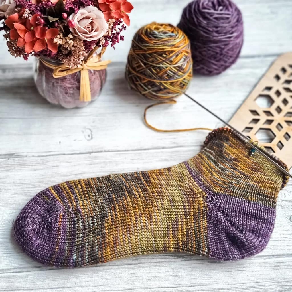 A almost finished knit sock laying flat next to some flowers