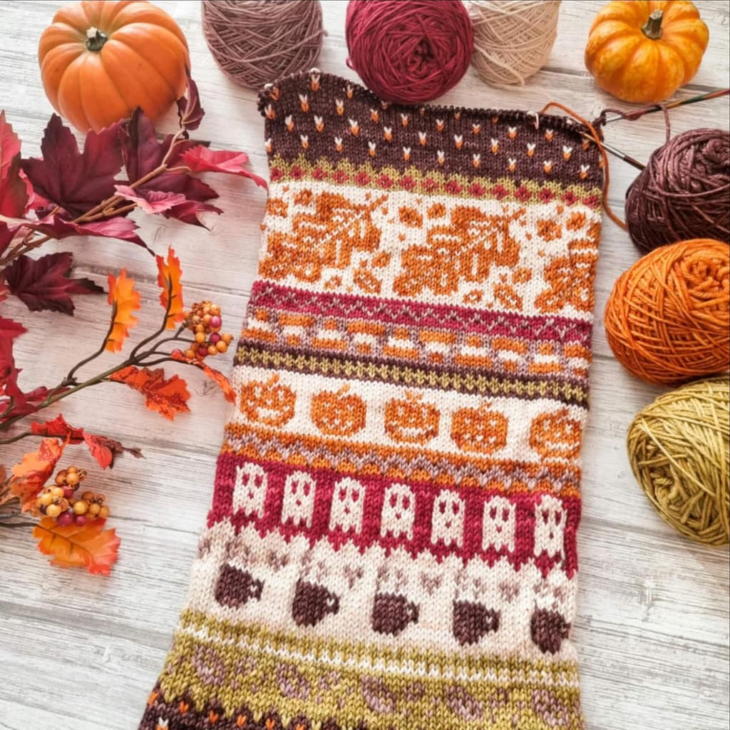 A knitted cowl in fall colors with many fall motives, surrounded by yarn and pumpkins