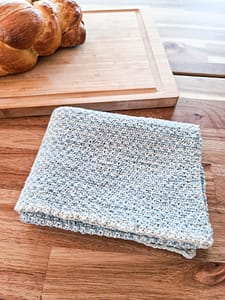 Cottage tea towel folded laying next to a wooden cutting board with bread on it