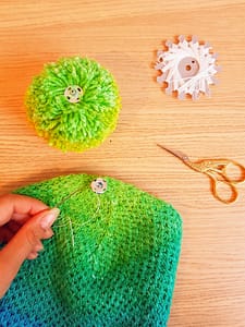 Top of a knitted hat, a pom-pom, and scissors, and a hand holding a threaded needle.