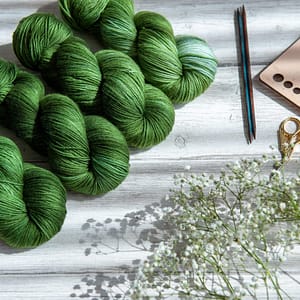 Four skeins of yarn in varying shades of green laying next to some flowers and knitting notions