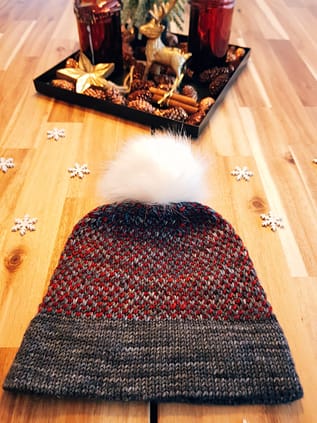 Two Tone Toque knit in red and grey laid flat with a white pom-pom. Winter decorations are visible in the background.