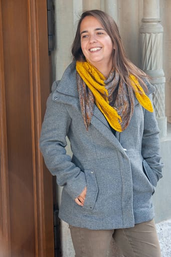 Woman wearing the two-colored Edge Of Winter Shawl and a grey coat, leaning against a wall.