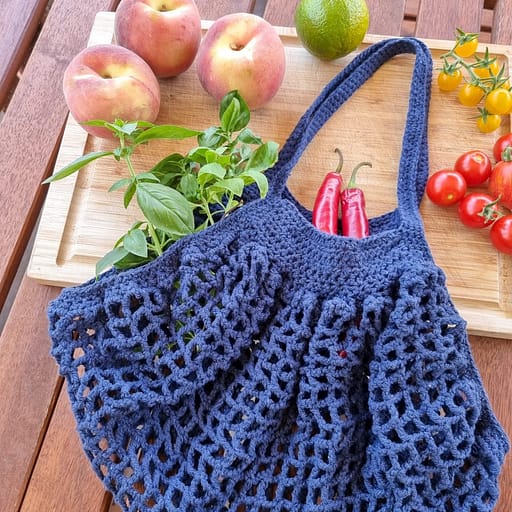 A crochet blue market bag with some fruits and vegetables around it