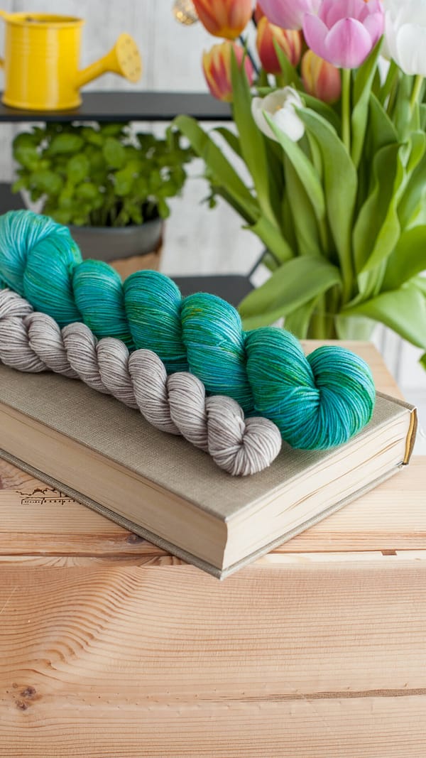 A yarn sock set in the colors Pond and Pebbles laying on top of a book