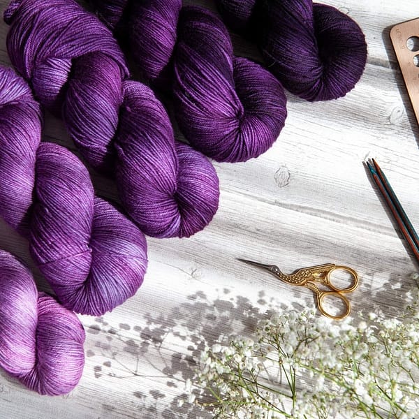 Five skeins of yarn in varying shades of purple laying next to some flowers and knitting notions