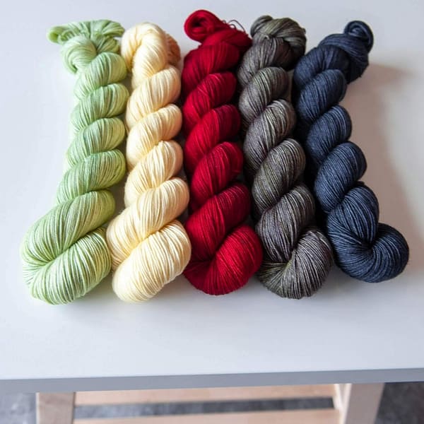 five skeins of yarn next to each other in the following colors: light green, vanilla, red, grey, and dark blue
