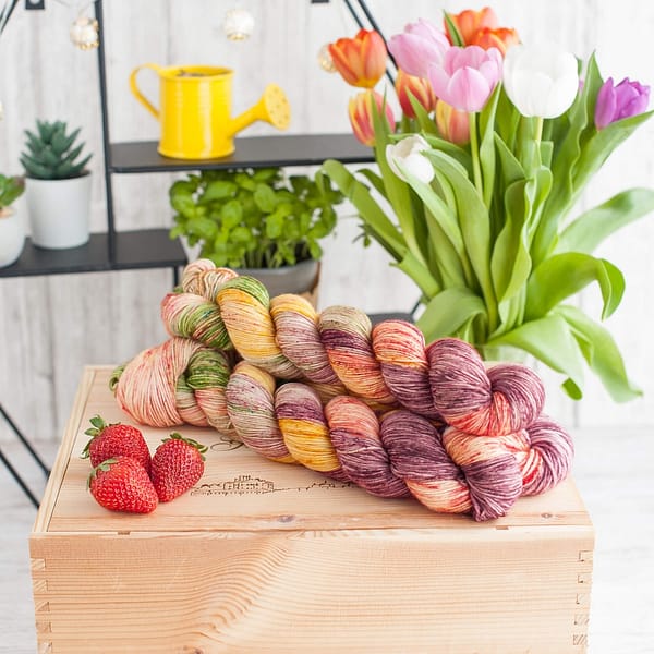 Three skeins of multicolored yarn with purple, green, yellow, and pink areas, with some flowers and strawberries in the background