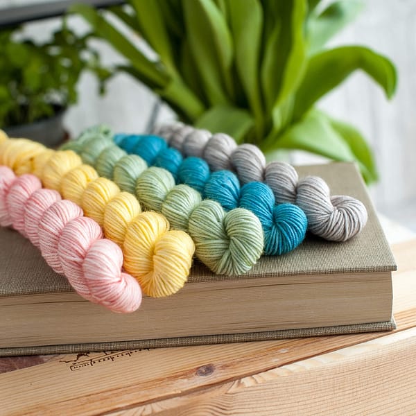 Five mini skeins of yarn in pastel blue, yellow, pink, green, and grey laying on top of a book
