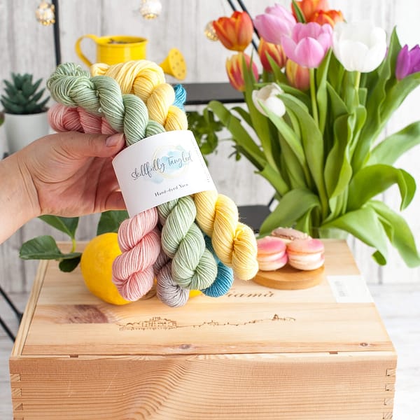 A hand holding five mini skeins of yarn in pastel blue, yellow, pink, green, and grey