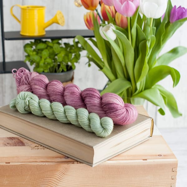 A yarn sock set in the colors Vintage Rose and Fresh Cut Grass laying on top of a book