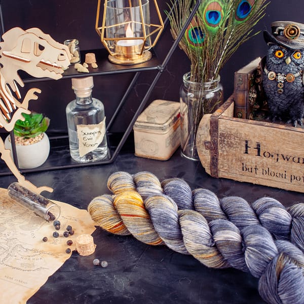 two skeins of grey yarn with yellow speckles laying in front of a wooden box and a wooden dinosaur
