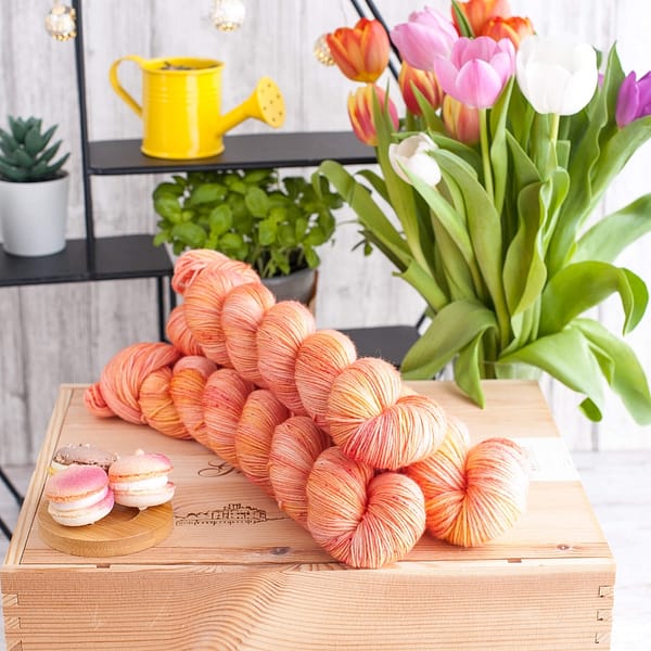Three skeins of peach colored yarn (yellow and pink),  on top of a book with some macarons and flowers