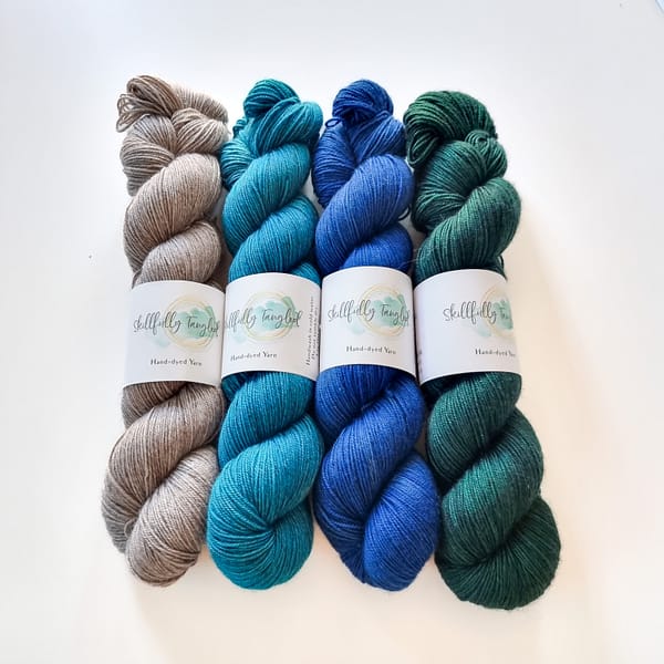 Four skeins of yak hand dyed yarn in grey, light blue, dark blue, and green. They are a kit for the Geogradient MKAL