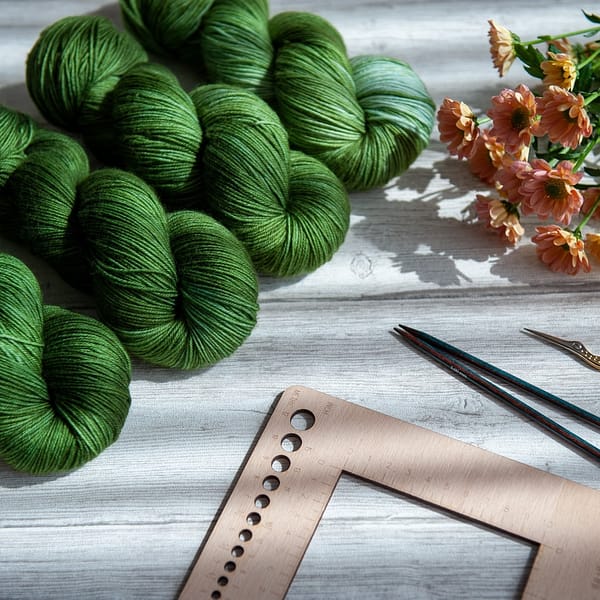 Four skeins of yarn in varying shades of green laying next to some flowers and knitting notions