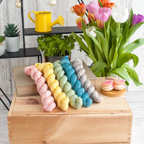 Five mini skeins of yarn in pastel blue, yellow, pink, green, and grey with flowers in the background